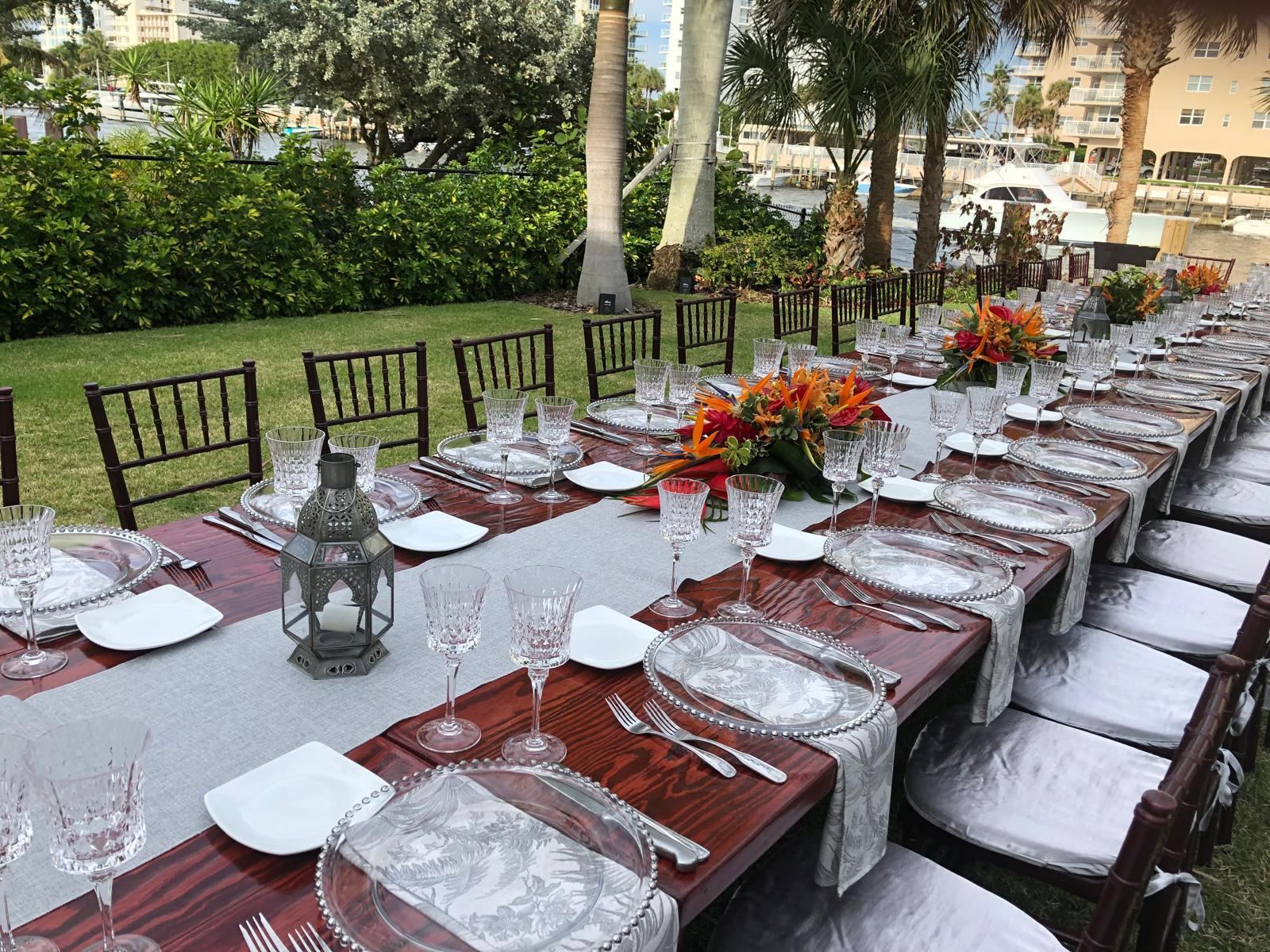 Long outdoor wedding table with crystal glassware and tropical flowers beside the water